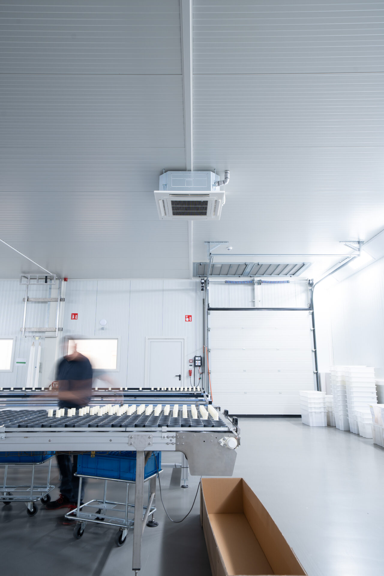 Industrial packing area with a conveyor belt carrying small white items; blurred worker on left, large doorway and ceiling-mounted HVAC unit overhead, and stacked crates on the right.