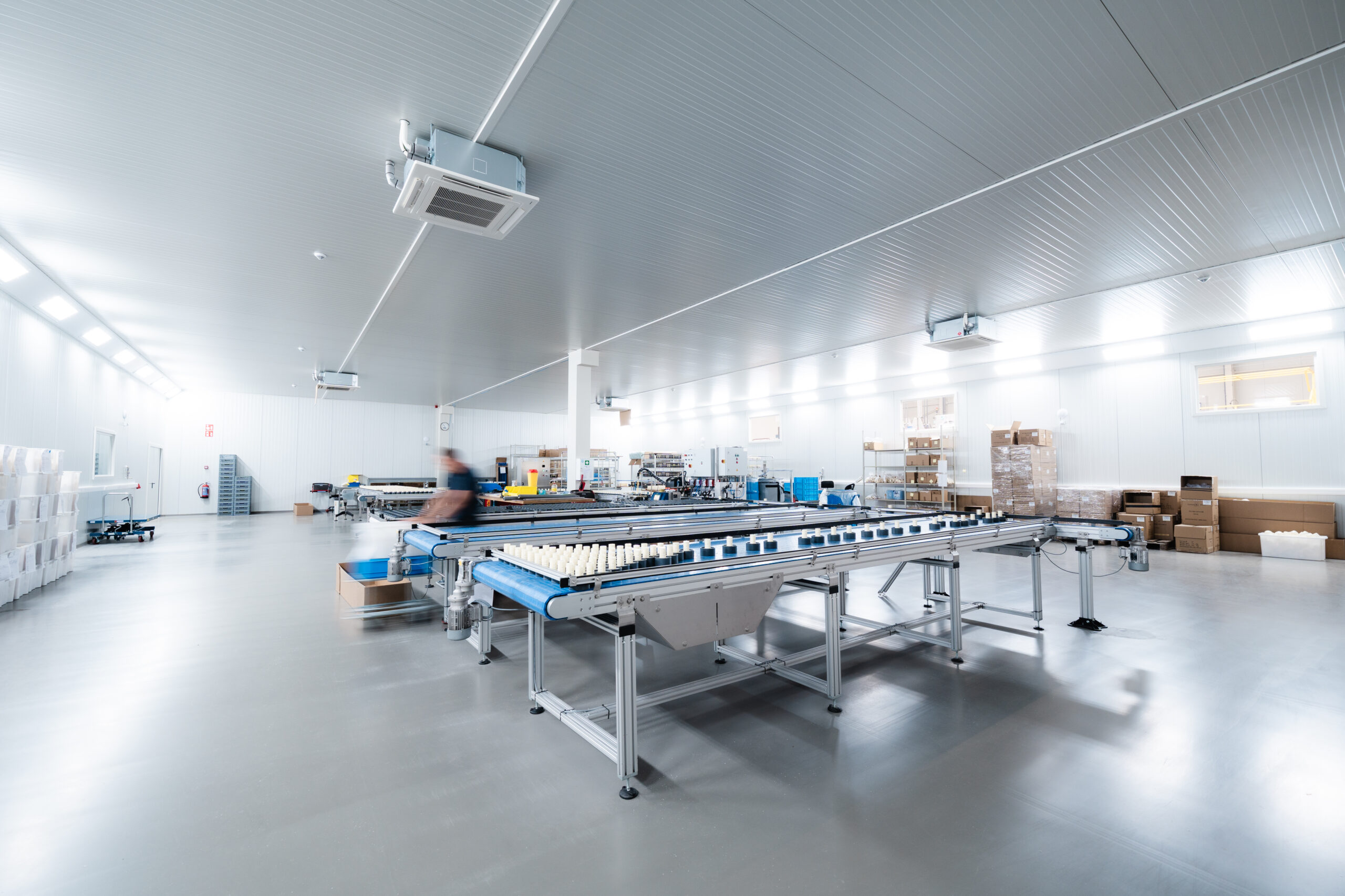 Clean, well-lit industrial processing hall with multiple conveyer belts and white bottles moving along blue rollers in a sterile facility.