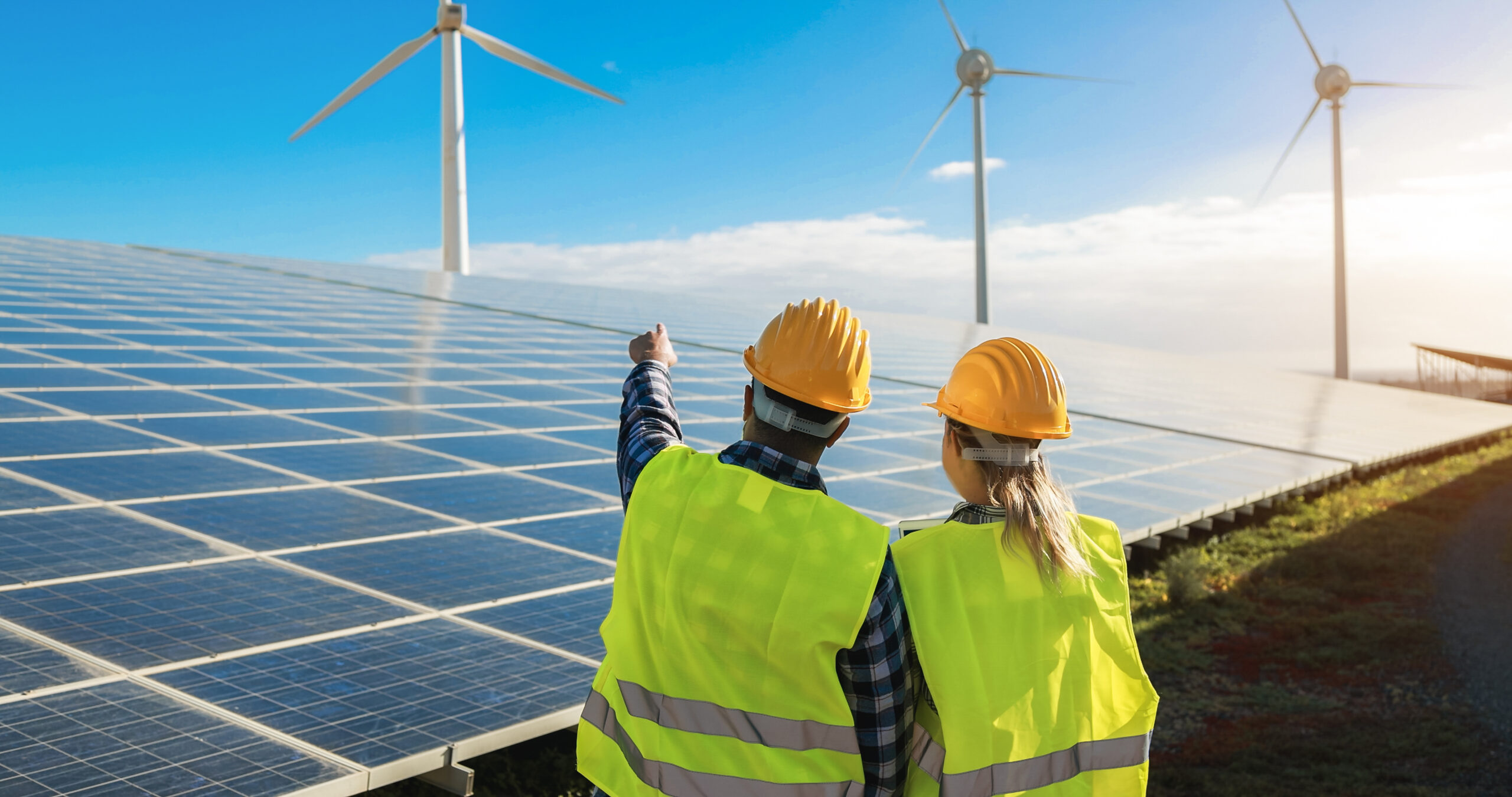 Two workers in yellow hard hats and high-visibility vests inspect solar panels at a solar farm with wind turbines in the background.