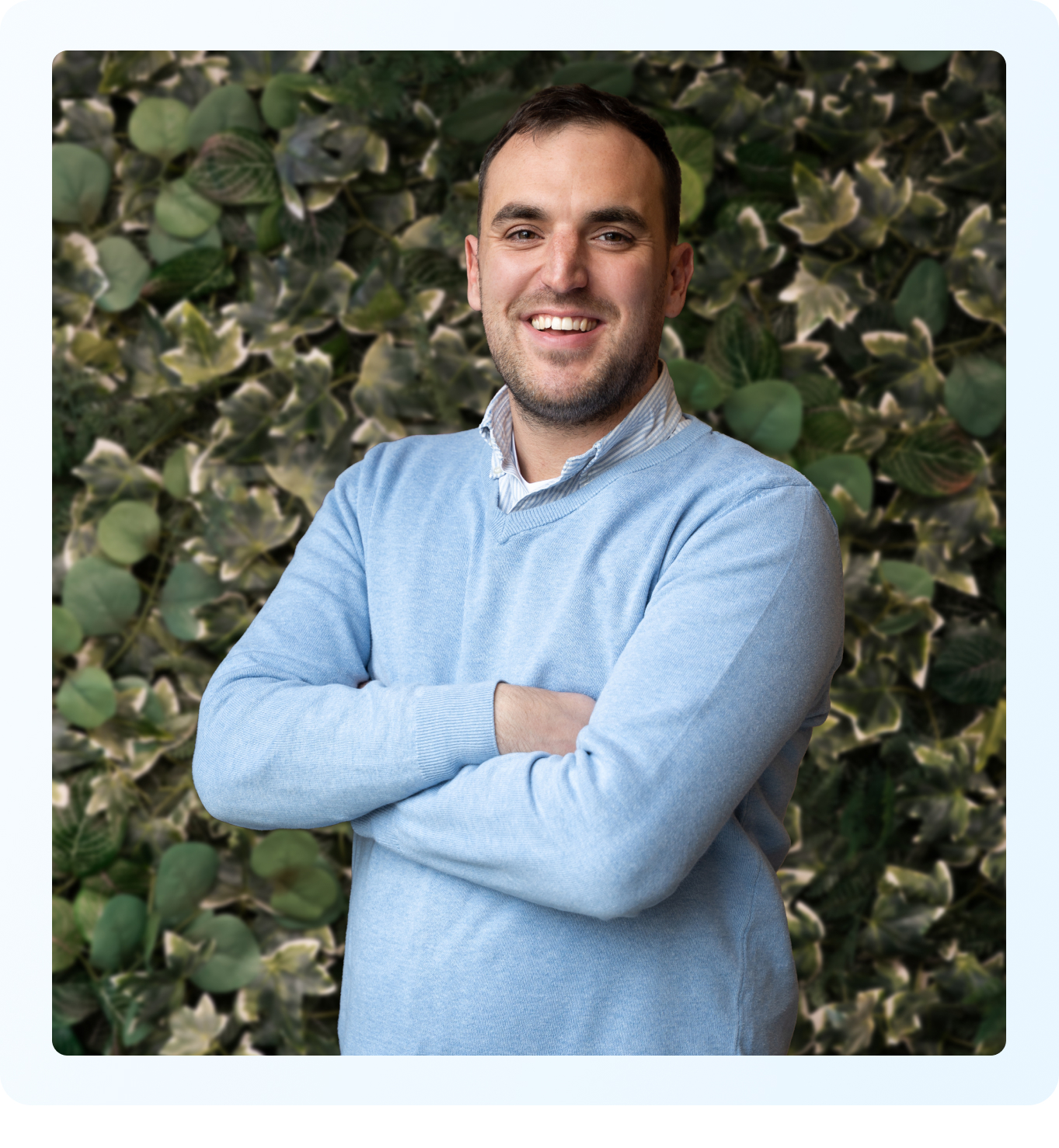 Smiling man with arms crossed in a light blue sweater, standing before green leafy plants.