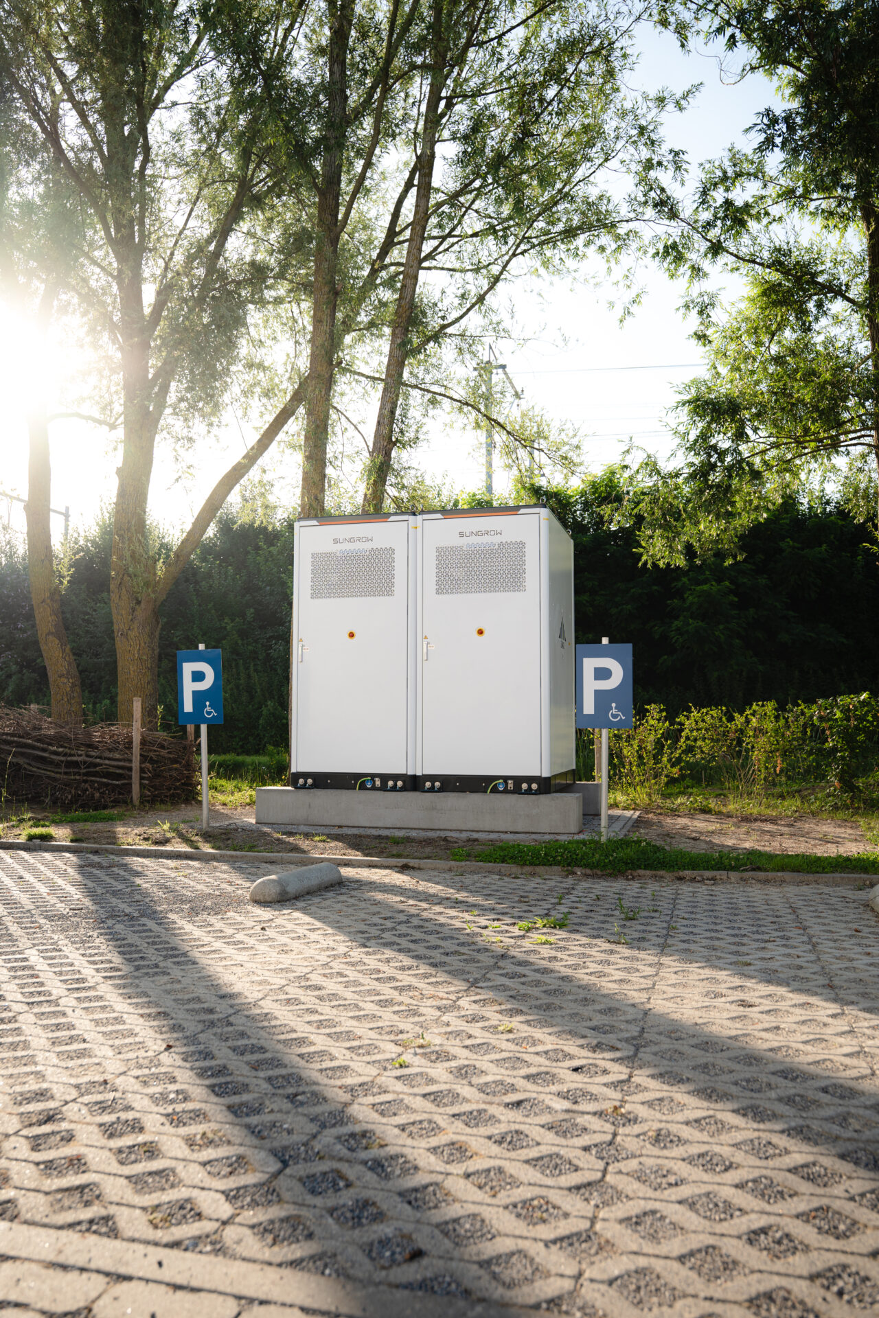 Two white Sungrow electrical cabinets on a concrete pad in a parking area, flanked by handicap signs, with trees in the background.