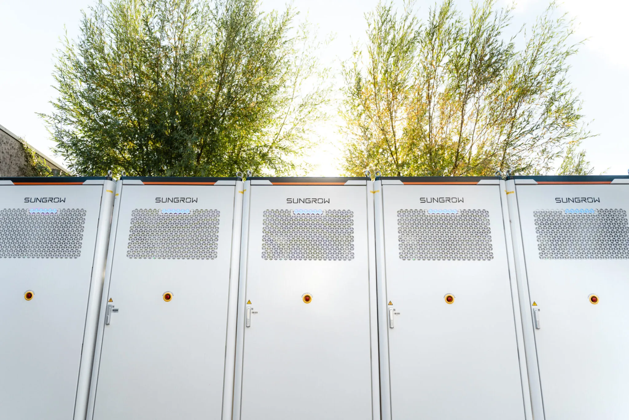 Row of white Sungrow equipment cabinets with vented panels, outdoors against trees.
