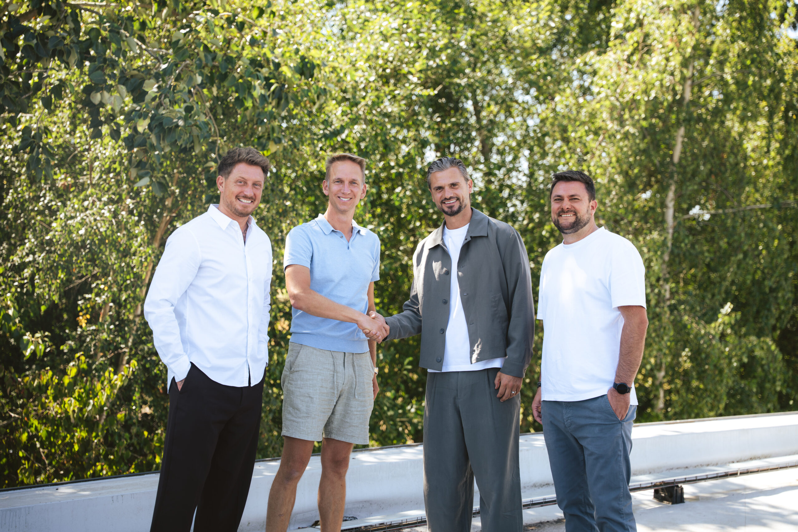 Four men stand outdoors on a sunny day, shaking hands in front of greenery and a white railing behind them.