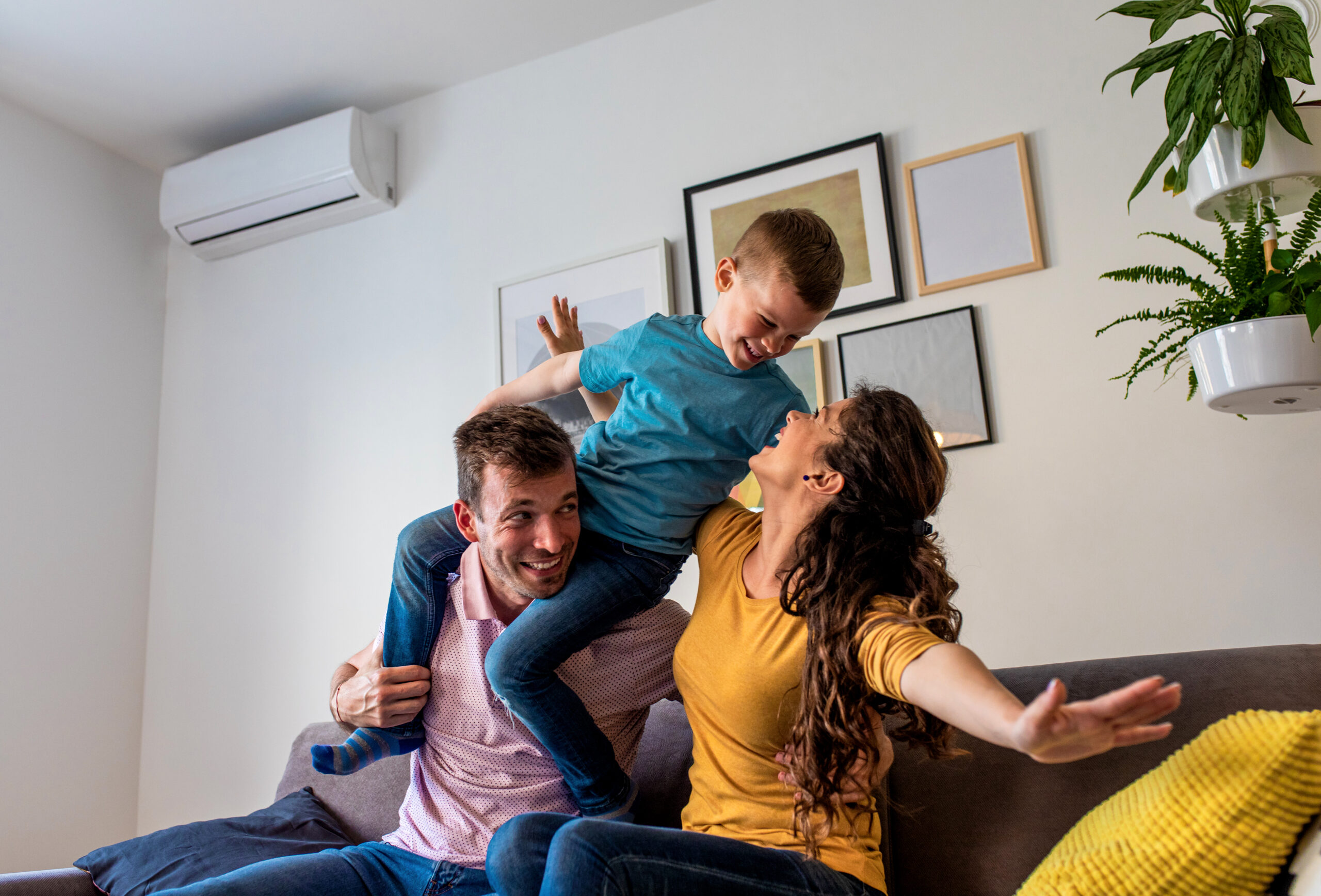 Father and mother lift their smiling son up in the air on a sofa in a bright living room. Framed art on the wall and potted plants visible behind them.