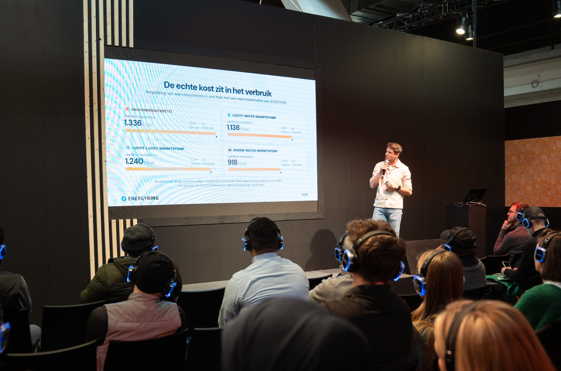 A speaker presenting a slide titled "The Real Cost Is in Consumption" to a seated audience on a conference stage, with a large screen displaying charts and attendees wearing in-ear headphones.